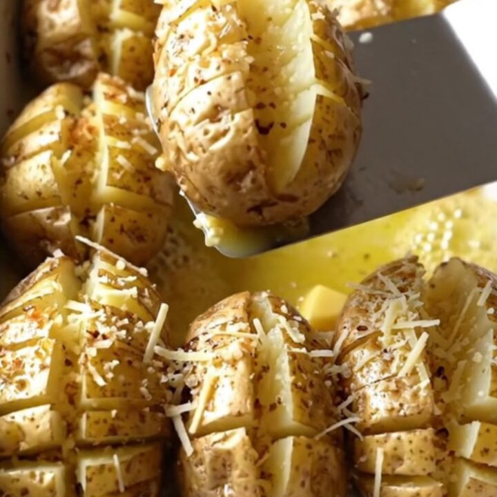 A top-down view of raw, accordion-cut russet potatoes in a white ceramic dish being drizzled with garlic butter.