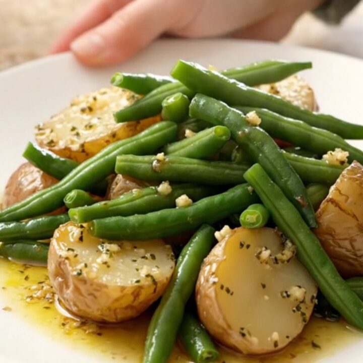 A close-up view of tender green beans and halved baby red potatoes in a garlic butter sauce inside a slow cooker.