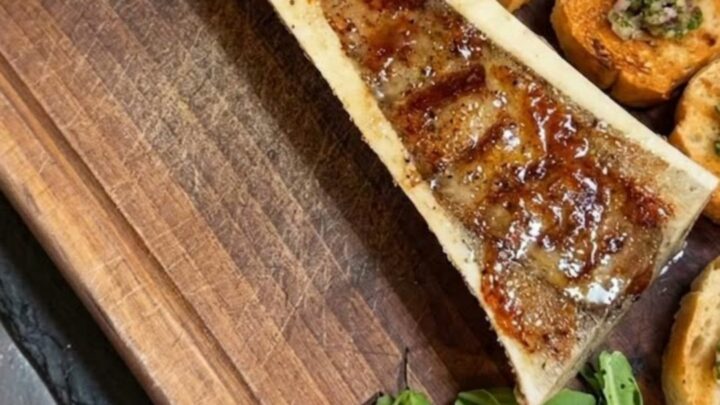 A close-up of hot, bubbling roasted beef bone marrow on a wooden plank with crostini.