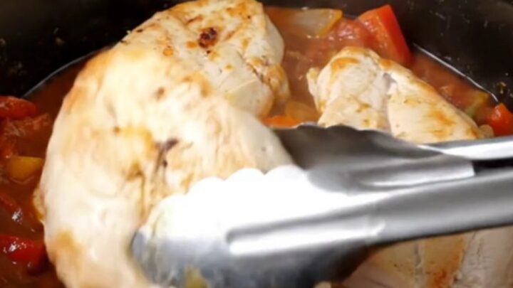 A top-down view of raw chicken breasts, crushed tomatoes, bell peppers, and onions in a slow cooker insert.