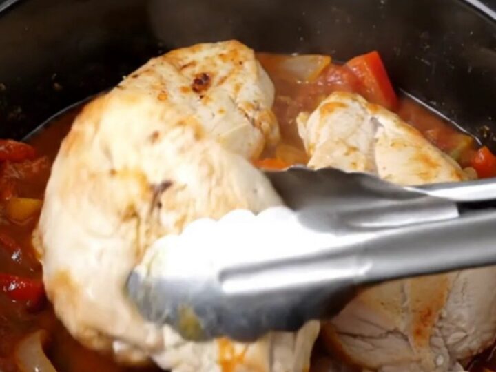 A top-down view of raw chicken breasts, crushed tomatoes, bell peppers, and onions in a slow cooker insert.