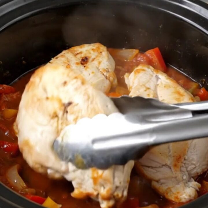 A top-down view of raw chicken breasts, crushed tomatoes, bell peppers, and onions in a slow cooker insert.