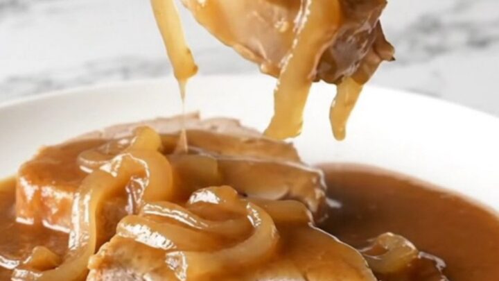 A close-up view of cooked pork steaks smothered in a rich brown onion gravy on a white plate.
