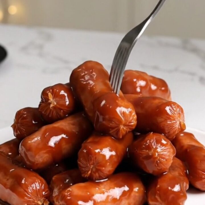 A close-up of plump mini smoked sausages coated in a thick, dark, and glossy barbecue glaze on a white plate.