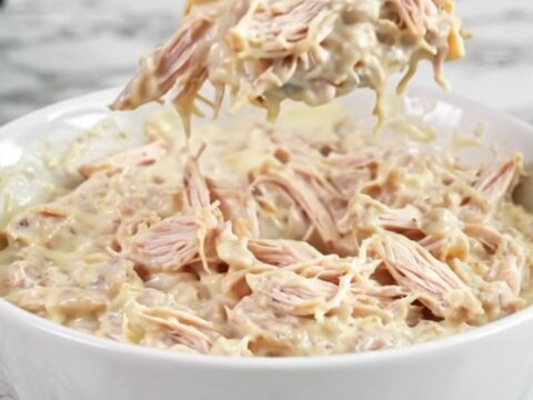 A close-up of creamy Texas Trash Chicken Dip in a white bowl with a spoon, showing a rich, melted cheese texture.
