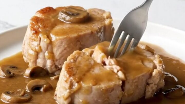 A close-up view of tender, cooked pork loin chops covered in a rich brown gravy with mushrooms and bacon pieces.