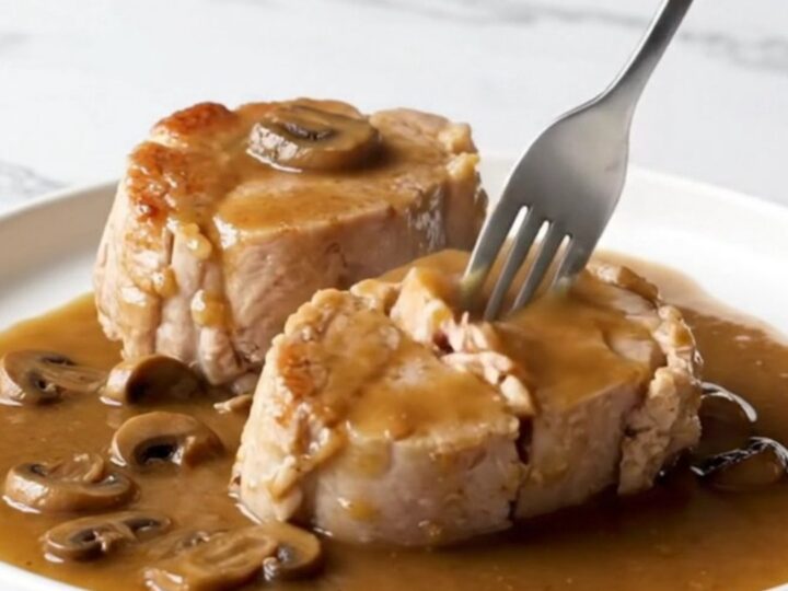 A close-up view of tender, cooked pork loin chops covered in a rich brown gravy with mushrooms and bacon pieces.