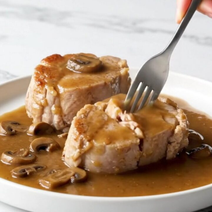 A close-up view of tender, cooked pork loin chops covered in a rich brown gravy with mushrooms and bacon pieces.