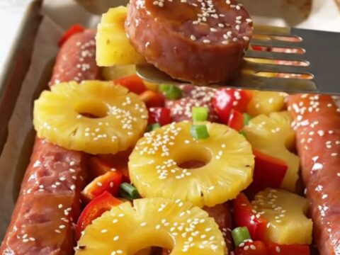 A sheet pan filled with browned kielbasa coins, golden roasted pineapple, and red bell peppers garnished with sesame seeds.