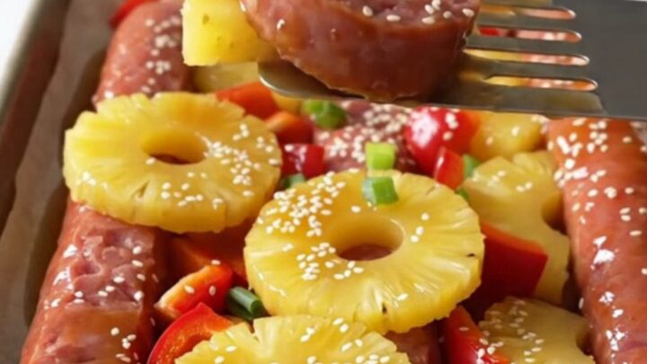 A sheet pan filled with browned kielbasa coins, golden roasted pineapple, and red bell peppers garnished with sesame seeds.