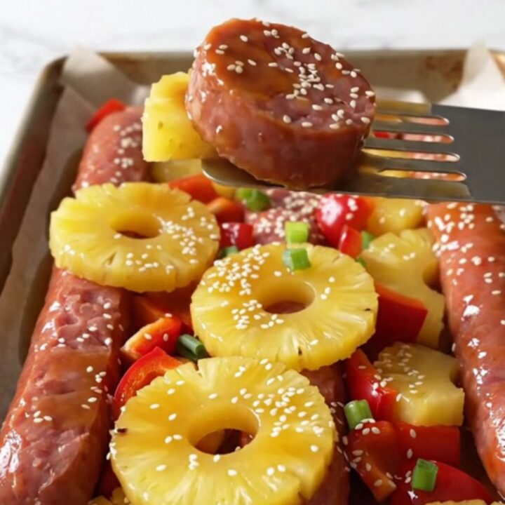 A sheet pan filled with browned kielbasa coins, golden roasted pineapple, and red bell peppers garnished with sesame seeds.