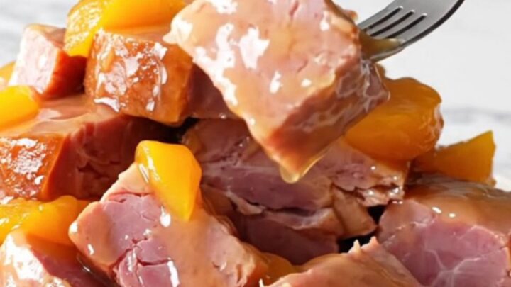 A close-up shot of slow-cooked ham chunks coated in a glossy peach and jalapeño glaze on a white marble surface.