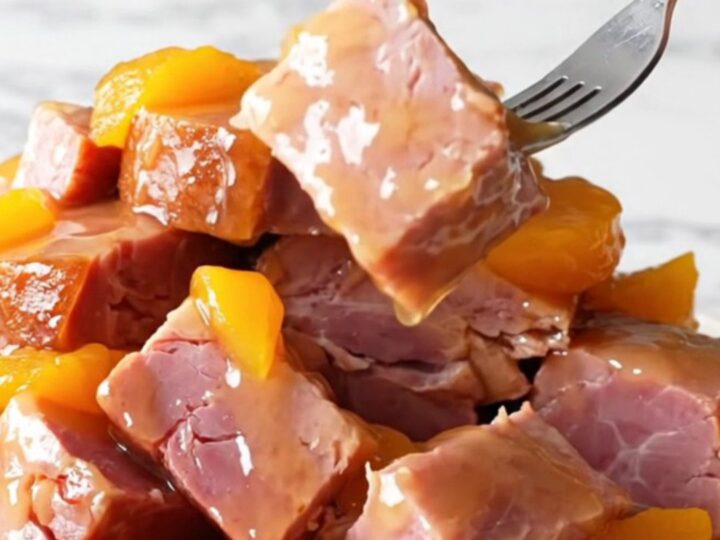 A close-up shot of slow-cooked ham chunks coated in a glossy peach and jalapeño glaze on a white marble surface.