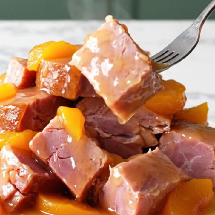 A close-up shot of slow-cooked ham chunks coated in a glossy peach and jalapeño glaze on a white marble surface.