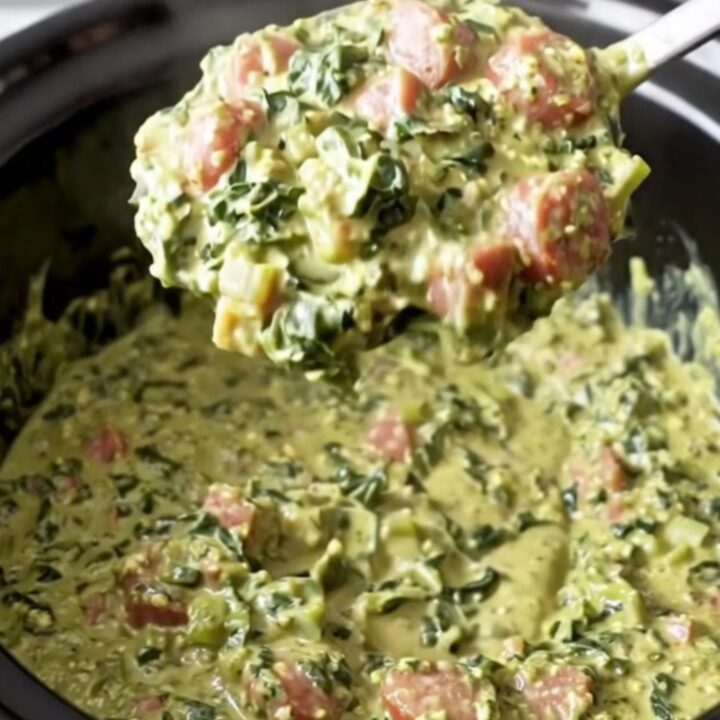 A close-up view of the creamy Southern swamp dip in a black slow cooker insert, showing bits of sausage and greens.