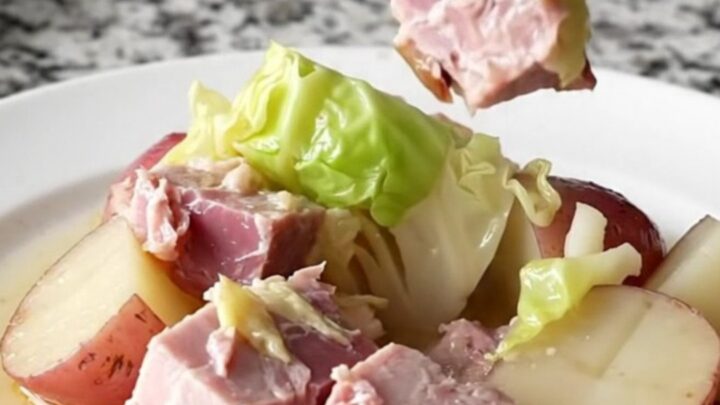 A close-up of a slow cooker filled with large pink ham chunks, green cabbage wedges, and red potatoes in a light broth.