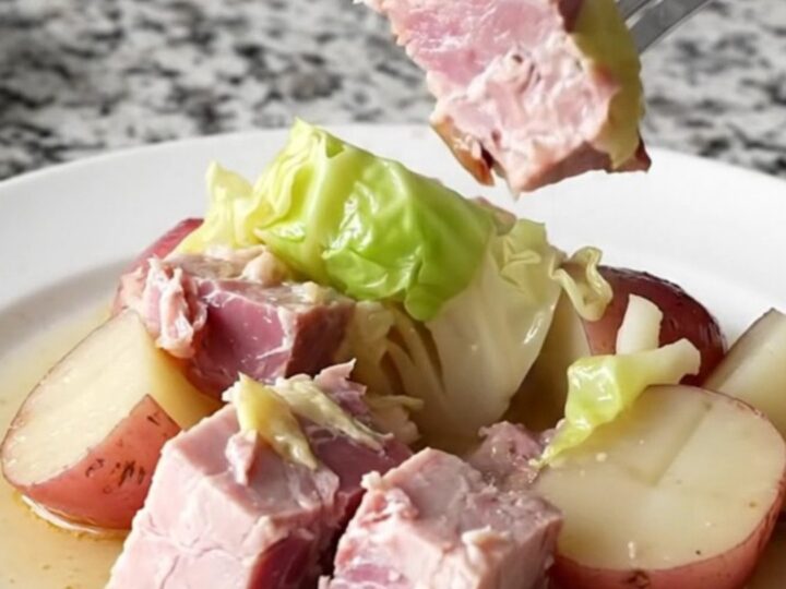 A close-up of a slow cooker filled with large pink ham chunks, green cabbage wedges, and red potatoes in a light broth.