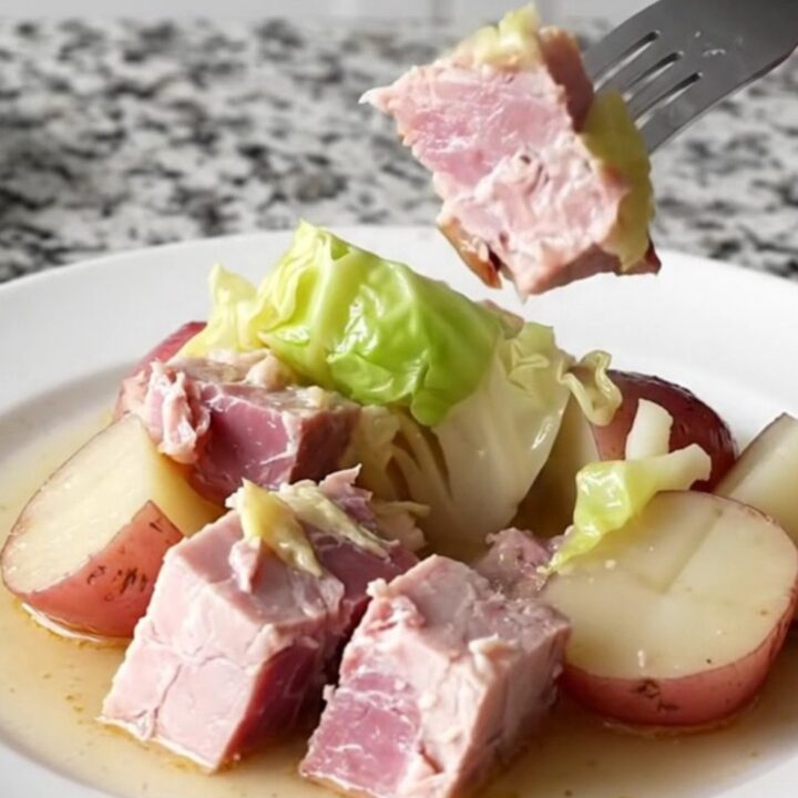 A close-up of a slow cooker filled with large pink ham chunks, green cabbage wedges, and red potatoes in a light broth.