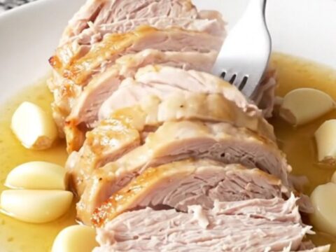 A beautifully cooked, golden-tan turkey breast sitting in a black slow cooker insert with aromatic garlic and herbs.