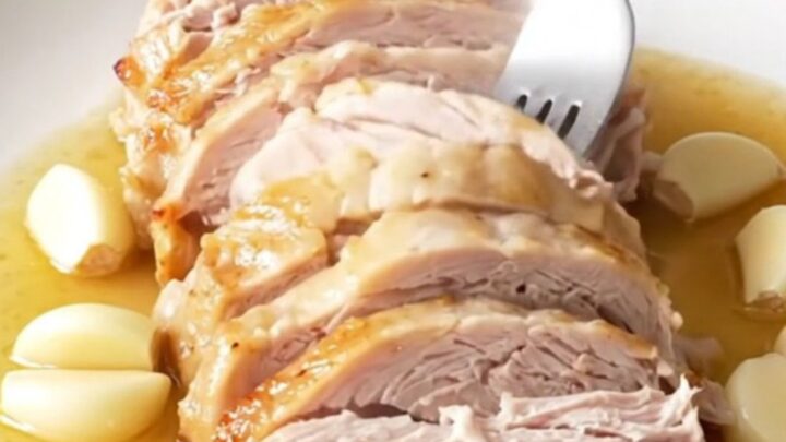 A beautifully cooked, golden-tan turkey breast sitting in a black slow cooker insert with aromatic garlic and herbs.