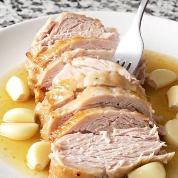 A beautifully cooked, golden-tan turkey breast sitting in a black slow cooker insert with aromatic garlic and herbs.