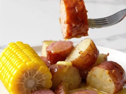 A slow cooker filled with sliced kielbasa, corn cobs, and red potatoes ready to be cooked.