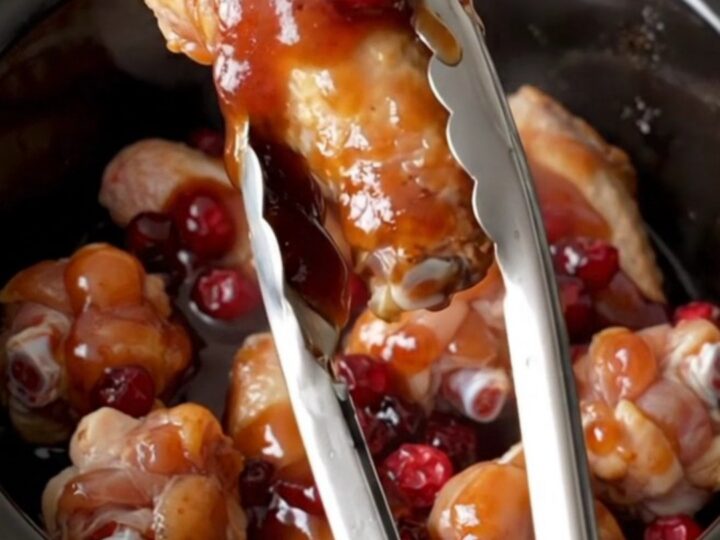 A close-up of tender, glazed chicken wings in a slow cooker, featuring dark red cranberry hoisin sauce and soft cranberries.