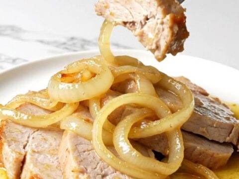 A close-up shot of juicy slow-cooked pork chops topped with soft white onions in a golden mojo sauce.