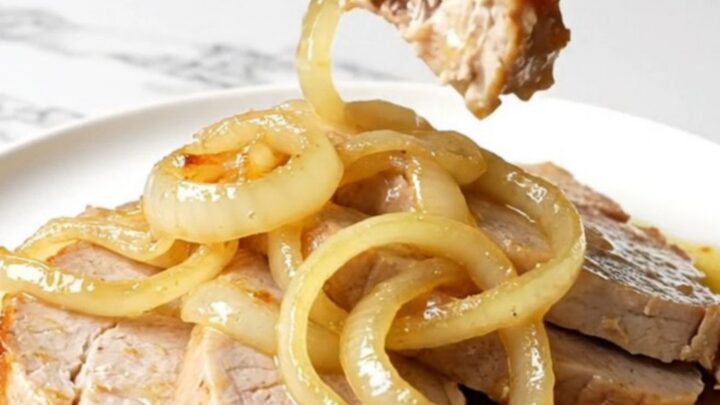 A close-up shot of juicy slow-cooked pork chops topped with soft white onions in a golden mojo sauce.
