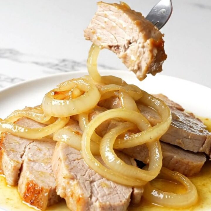 A close-up shot of juicy slow-cooked pork chops topped with soft white onions in a golden mojo sauce.