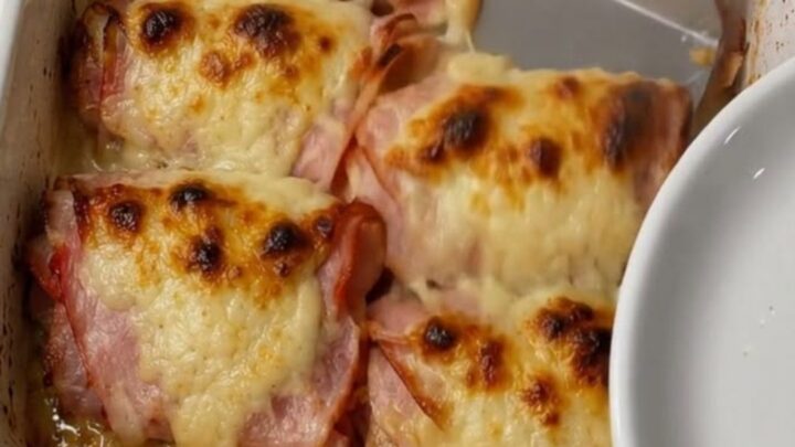 A white ceramic baking dish filled with six golden-brown, cheesy ham rolls on a granite counter.