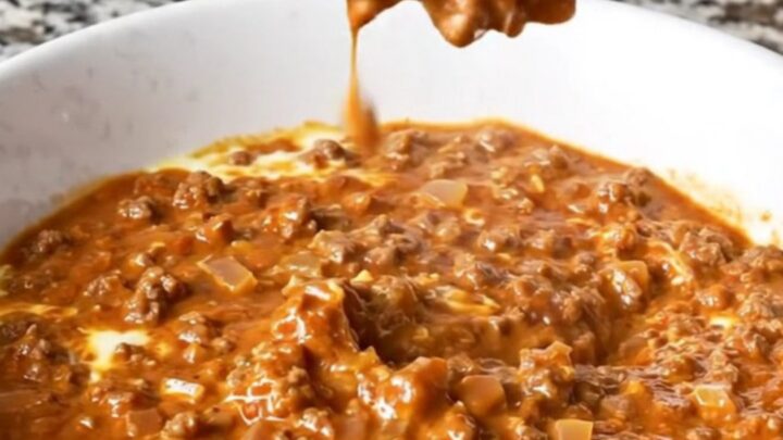 A warm, creamy reddish-brown Cincinnati chili dip in a white bowl with beef crumbles and visible spices.