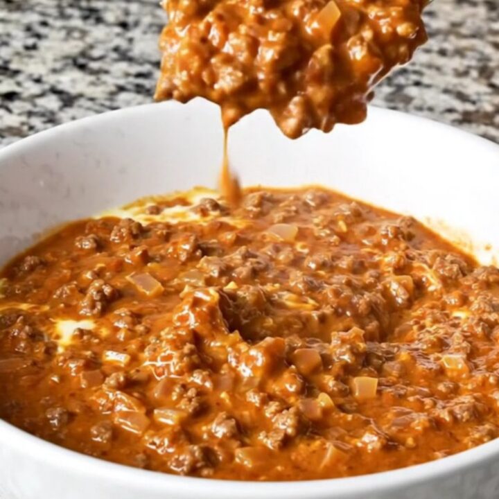 A warm, creamy reddish-brown Cincinnati chili dip in a white bowl with beef crumbles and visible spices.