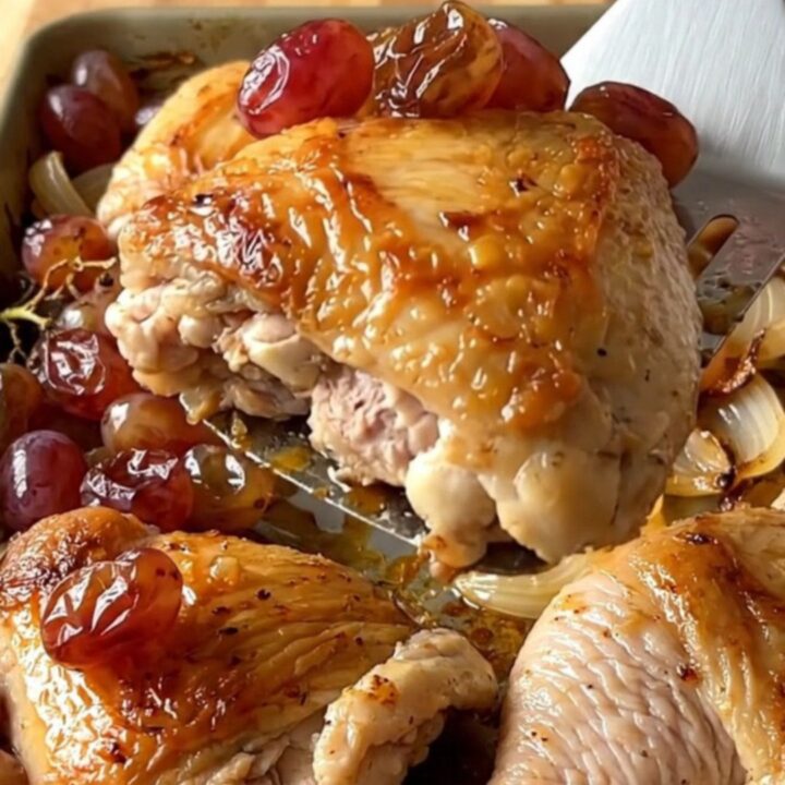 A top-down view of a sheet pan filled with roasted chicken thighs, red grapes, and red onion wedges.