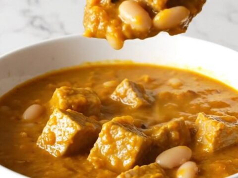 A bowl of hearty pork and pumpkin white bean chili with tender pork cubes and a thick orange sauce.