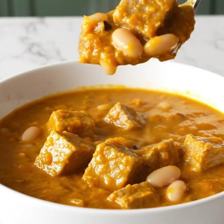 A bowl of hearty pork and pumpkin white bean chili with tender pork cubes and a thick orange sauce.