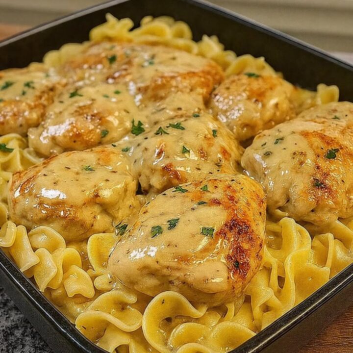 A square black pan filled with golden seared chicken cutlets resting over creamy, glossy egg noodles.