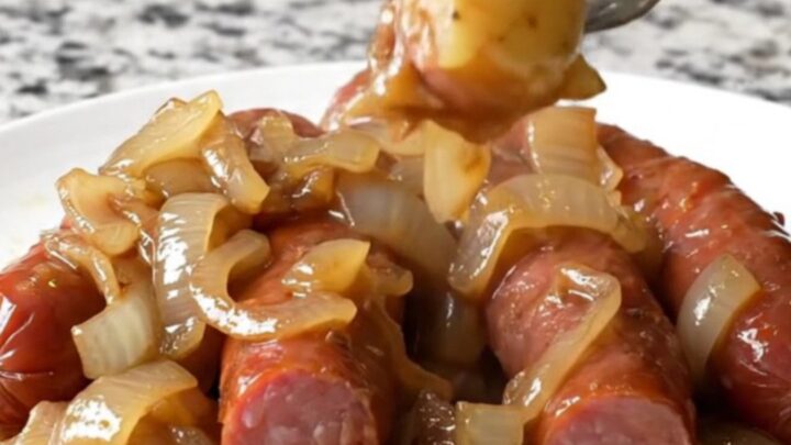A close-up of a white plate filled with sliced kielbasa, gold potatoes, and onions in a savory brown au jus.