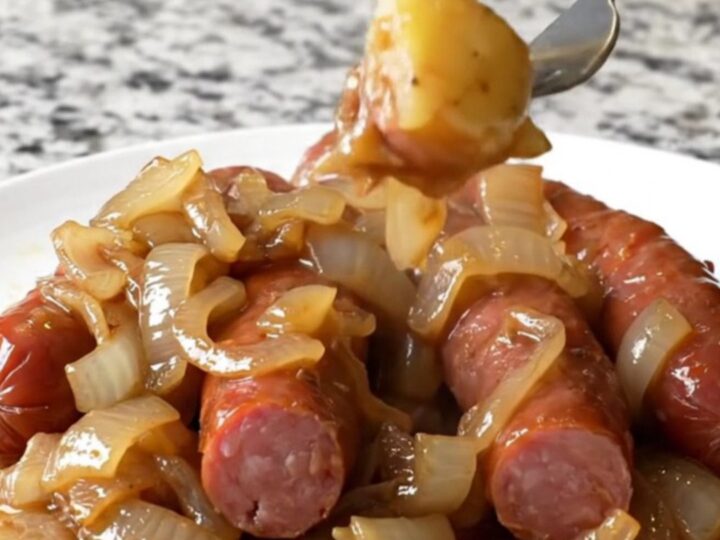 A close-up of a white plate filled with sliced kielbasa, gold potatoes, and onions in a savory brown au jus.