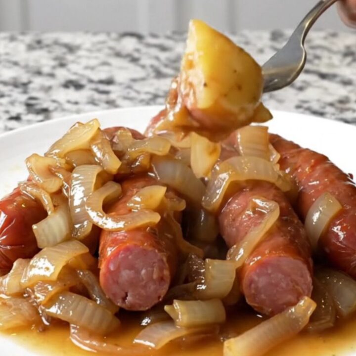 A close-up of a white plate filled with sliced kielbasa, gold potatoes, and onions in a savory brown au jus.