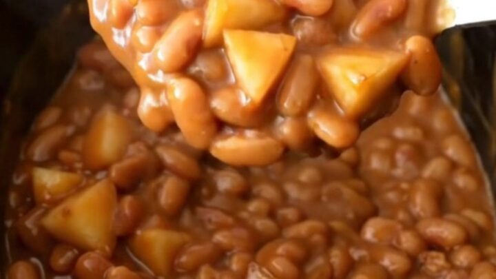 A close-up view of slow cooker apple bacon baked beans in a black stoneware insert, showing a rich brown texture.