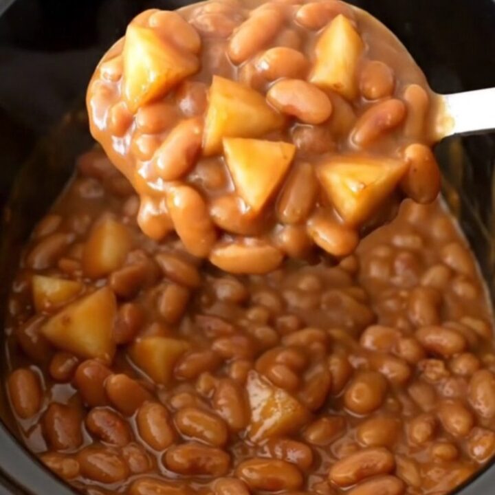 A close-up view of slow cooker apple bacon baked beans in a black stoneware insert, showing a rich brown texture.