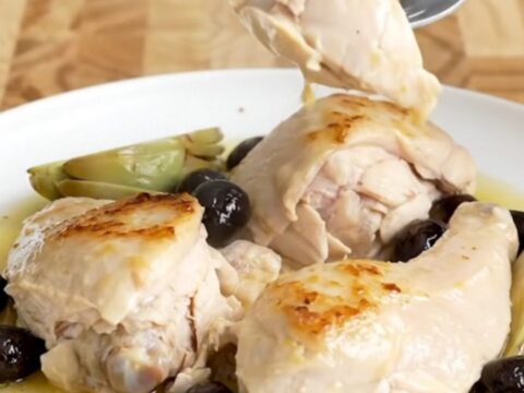 A slow cooker filled with chicken breasts, artichokes, and olives in a light lemon sauce on a butcher-block counter.