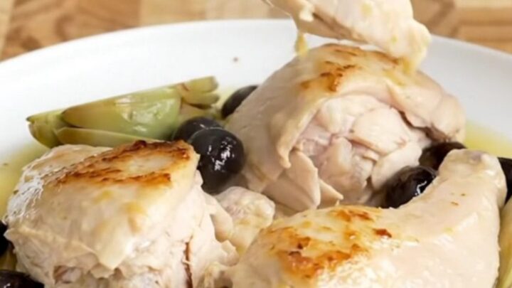 A slow cooker filled with chicken breasts, artichokes, and olives in a light lemon sauce on a butcher-block counter.