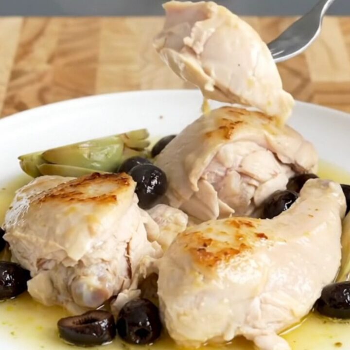 A slow cooker filled with chicken breasts, artichokes, and olives in a light lemon sauce on a butcher-block counter.