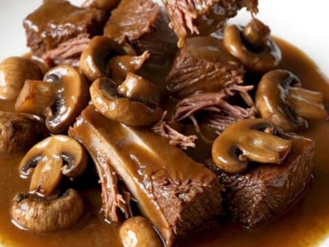 A close-up of tender brown beef tips and whole mushrooms in a rich gravy inside a black slow cooker.