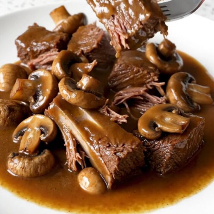 A close-up of tender brown beef tips and whole mushrooms in a rich gravy inside a black slow cooker.