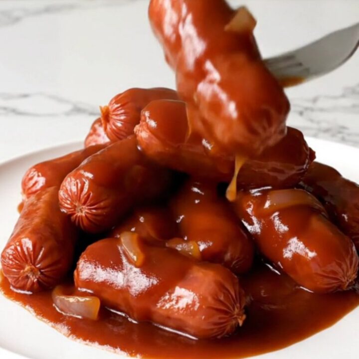 A slow cooker filled with smoked cocktail sausages in a rich, dark red barbecue sauce with visible onion rings.