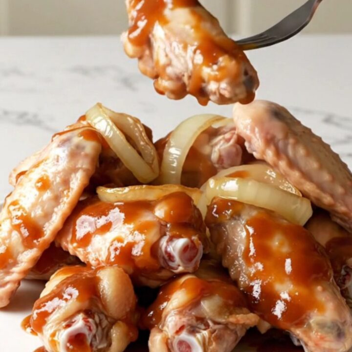 A plate of tender slow-cooked BBQ chicken wings coated in a dark, glossy sauce with soft onion rings.