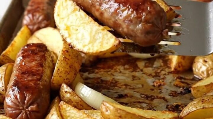 A sheet pan filled with golden-brown bratwurst, crispy potato wedges, and soft onions.